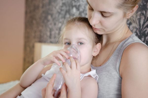 Bambina bionda, si tiene la mascherina di aerosol sul viso, è seduta sulle ginocchia della mamma che la aiuta a fare l'aerosol per trattare la tosse grassa